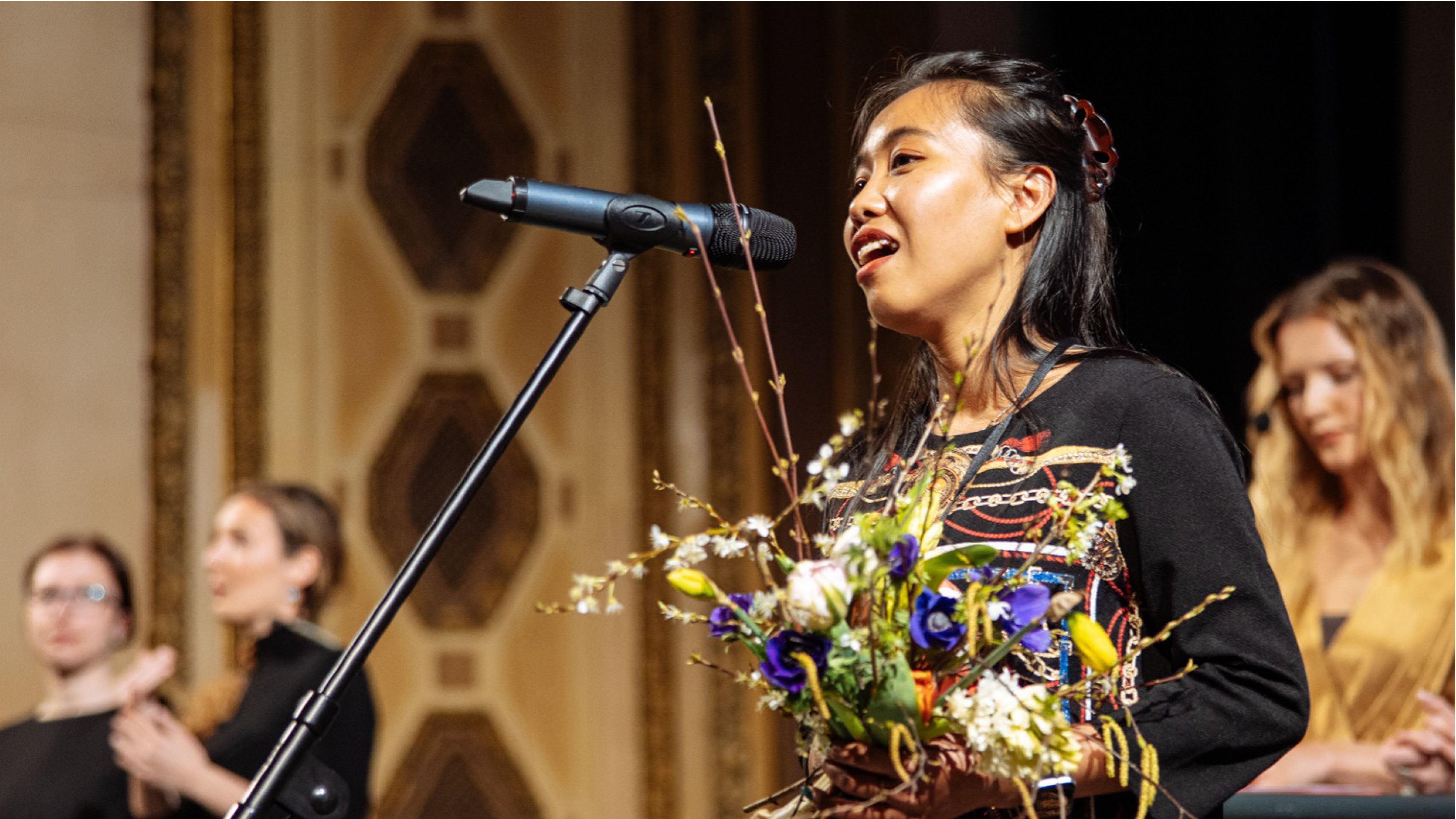 Na pódiu stojí režisérka Snow Hnin Ei Hlaing, drží kytici jarních květin a hovoří do mikrofonu při závěrečné ceromonii.