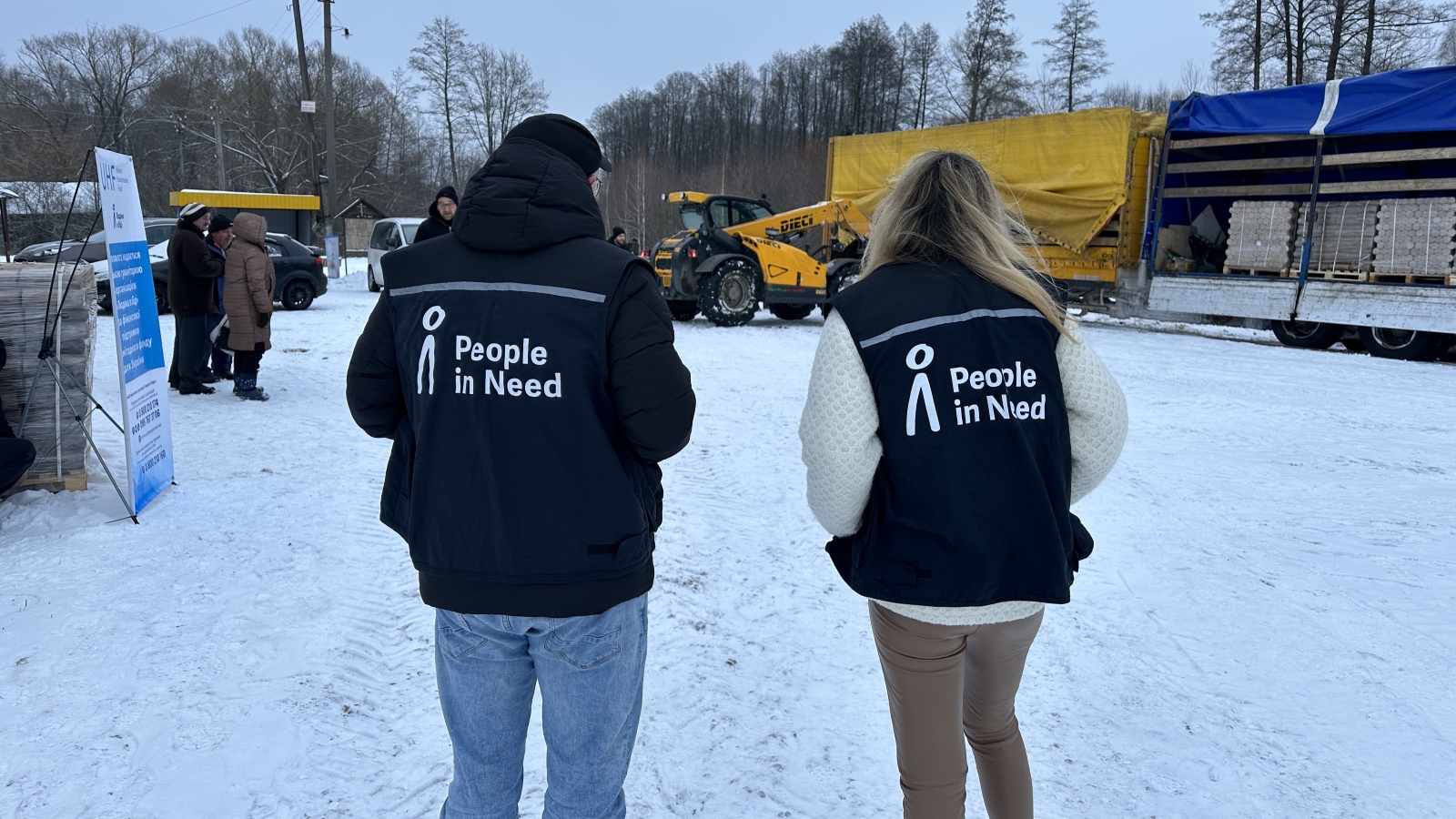 Dvě osoby s vestami s logem Člověk v tísni stojí venku na zasněžené silnici a hledí na nákladní auto a bagr.