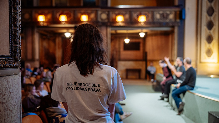 Dotazník Osoba s delšími vlasy stojí zády k fotoaparátu uprostřed sálu, na sobě má bílé tričko s nápisem „Moje srdce bije pro lidská práva“. Před ní je pódium, kde sedí několik lidí zapojených do diskuse, naproti nim sedí publikum.