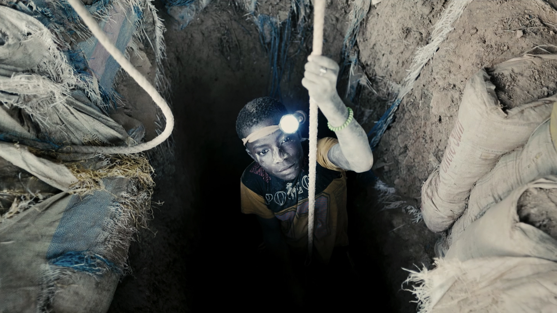 Mladý africký chlapec s čelovkou na hlavě se na laně spouští do hluboké černé díry pro vodu. /A young African boy with a headlamp on his head descends on a rope into a deep black hole for water.